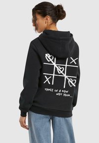 Felpa con cappuccio nera realizzata in tessuto morbido, con un design di tris con cuori e croci bianchi sul retro. Il testo recita "TRE IN FILA NON TUO...".