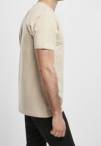 Muž v béžovém tričku s krátkým rukávem a černých kalhotách stojící na jednobarevném světle šedém pozadí, boční pohled.