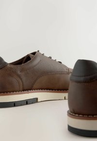 Zapatos de cuero marrón con un upper texturizado, una puntera lisa y una suela blanca contrastante con acentos negros. Los cordones son grises.