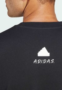 T-shirt en coton noir avec un logo Adidas blanc et le texte "A.D.I.D.A.S." dans le haut du dos. Col ras du cou, manches courtes, tissu texturé.