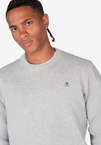 Suéter gris de punto con cuello redondo, tejido texturizado y un pequeño logo de calavera y tibias cruzadas en el pecho.