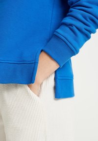 Blue sweatshirt featuring ribbed cuffs and hem, paired with ribbed white pants. Pocket visible on the right side. Soft, textured fabric.