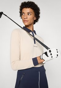 Beige long-sleeve polo with navy collar accents, paired with navy golf pants. White golf glove features black grip details. Golf club across shoulder.