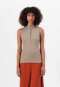 Woman with curly hair wearing a sleeveless beige button-up top and loose rust-colored pants standing against a white background.