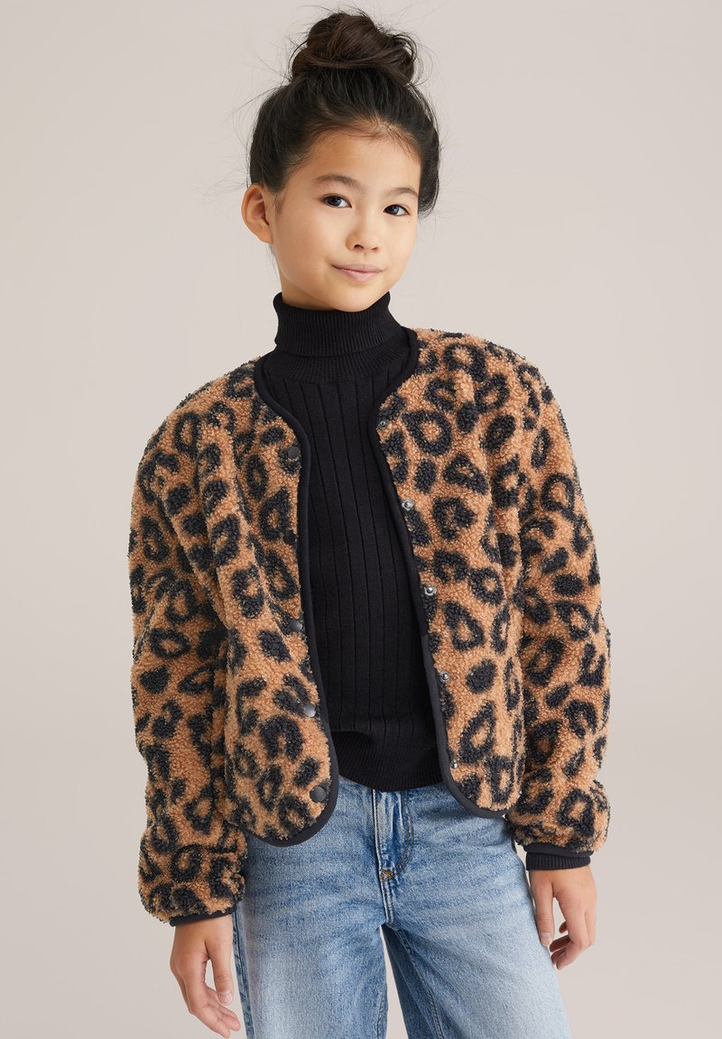 Giacca in pelliccia sintetica con stampa leopardata e dettagli neri, colletto rotondo e chiusure a scatto. Indossata sopra un dolcevita nero a costine e jeans.