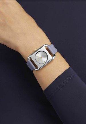 Montre rectangulaire argentée avec un cadran blanc texturé, des accents dorés et un bracelet en cuir violet clair, présentée sur un poignet.
