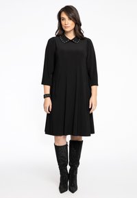 Schwarzes Kleid mit rundem Kragen, der mit Nieten verziert ist, 3/4-Ärmeln, plissiertem Rock und zwei Seitentaschen. Kombiniert mit knielangen, strukturierten schwarzen Stiefeln.