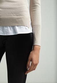 Pull beige en tricot avec ourlet côtelé porté sur une chemise blanche, associé à des leggings noirs. Gros plan sur le bras et la main reposant sur la jambe.