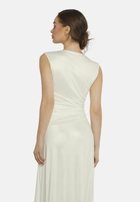 Mouwloze, crèmekleurige jurk met een getailleerde bodice en een vloeiende rok. Materiaal lijkt glad met een subtiele glans. Geplooide details bij de taille.
