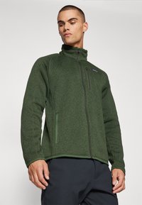 Dark green fleece jacket with high collar, full zip front, and fitted silhouette. Includes zippered pockets and textured fabric.