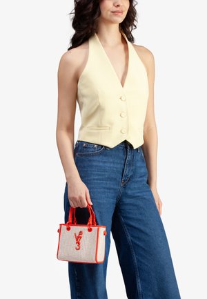 Donna che indossa un gilet senza maniche giallo pallido e jeans blu, mentre tiene una piccola borsa color crema e rosso con il logo "V73".