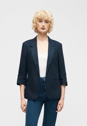 Vrouw met kort krullend blond haar, gekleed in een donkerblauw colbert over een wit shirt en een blauwe spijkerbroek, staand tegen een effen lichte achtergrond.