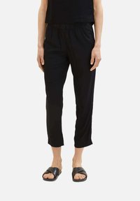 Zwarte cropped pantalon met een elastische tailleband, gladde textuur en lichte glans. Gecombineerd met zwarte slide sandalen.