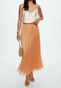 Plisseret, skinnende, orange maxi-skjørt med en høj-lav kant. Bæres med en krème farvet tanktop og accessoriseret med en vævet taske og strop sandaler.