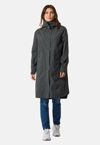 Cappotto impermeabile grigio con colletto alto e zip frontale, maniche lunghe con polsini regolabili e vestibilità ampia, abbinato a jeans blu e sneakers bianche.