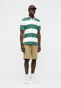 Άνδρας που φοράει πράσινη και λευκή ριγωτή μπλούζα πόλο, μπεζ βερμούδα μέχρι το γόνατο, λευκά αθλητικά παπούτσια, λευκές κάλτσες και καφέ μπέιζμπολ καπέλο, στέκεται όρθιος.