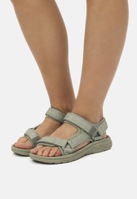 Sandalias verde claro con correas ajustables, acentos texturizados y una suela curvada para mayor comodidad. Diseño de punta abierta con detalles de hebilla visibles.