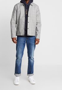 Chaqueta de cuadros gris con botones a presión sobre una sudadera azul marino, combinada con jeans azules y zapatillas blancas, ofreciendo un aspecto casual en capas.