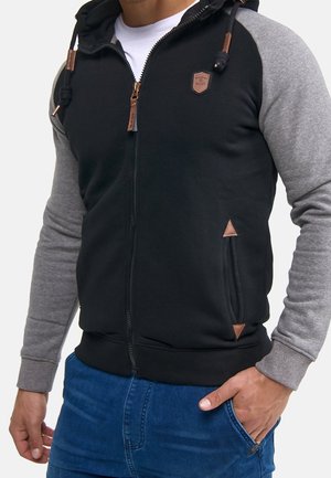 Sweat à capuche noir avec fermeture éclair, manches grises, tirettes en cuir marron, et une petite écusson sur la poitrine, porté par-dessus une chemise blanche et un jean bleu.