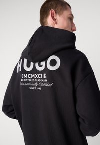 Černá mikina s kapucí, na přední straně s velkým bílým nápisem "HUGO" a složitým textem na zadní straně, vyrobená z měkké látky s uvolněným střihem a stahovací kapucí.