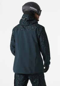 Ski jacket in dark teal with a patterned hood and upper back. Features smooth texture, waterproof material, and adjustable cuffs.