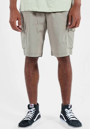Homme portant un short cargo beige et des baskets montantes noires avec des lacets blancs, debout devant un fond clair uni.