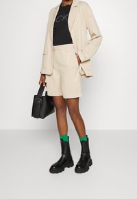 Beige blazer og shorts, sort t-shirt med logo, sorte chunky ankelstøvler, grønne sokker, holder en sort håndtaske. Glat og struktureret stof.