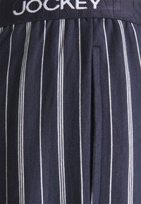 Mörkblå randiga shorts med vita vertikala linjer, med ett elastiskt midjeband märkt med "JOCKEY" och sidfickor. Mjuk tygtextur.