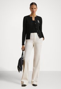 Μαύρη ζακέτα με πλεκτό σχέδιο, στρογγυλή λαιμόκοψη, χρυσό κουμπί στο κέντρο και κεντημένο λογότυπο. Συνδυασμένη με κρεμ παντελόνι φαρδύ στην ποδιά και τσάντα με πουά μοτίβο.