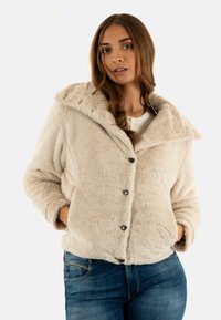 Chaqueta de piel sintética crema con un amplio cuello, cinco botones negros y dos bolsillos laterales, combinada con jeans azules. Textura suave y diseño cálido.