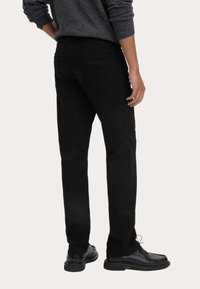 Černé kalhoty rovného střihu s hladkým povrchem, s zadními kapsami a jemnými švy, spárované s černými koženými botami.