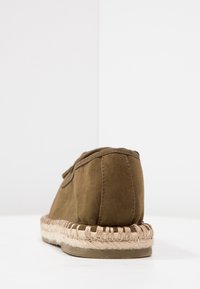 Braunes Wildleder-Schlupfschuh mit abgerundeter Ferse, ausgestattet mit einer Espadrille-artigen Jutesohle und dekorativen Nähten entlang der Kante.