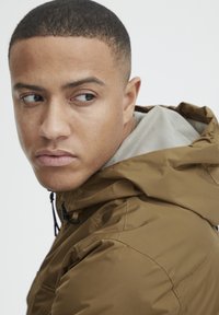 Brown hooded jacket with a textured gray inner lining, featuring a zipper and adjustable drawstrings, shown on a model from the side.