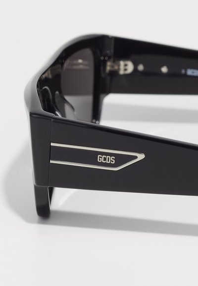 Ochelari de soare negri cu un cadru din plastic elegant, detalii argintii pe laterale și lentile închise la culoare. Prezintă un design unghiular și branding GCDS.
