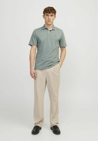 Zelené polo triko s límečkem, doplněné béžovými kalhotami a černými botami. Hladká látka bez viditelných vzorů či log.