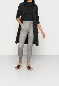 Svart trench coat över en svart topp, grå strukturerade joggers med dragsnöre, och svarta sandaler med bred rem. Minimalistiska designelement.