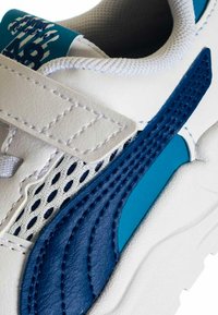 Scarpa da ginnastica bianca con dettagli blu, pelle testurizzata, inserti in mesh e chiusura con cinturino in Velcro; include una toppa decorativa con il logo sulla parte superiore.