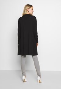 Melnās garās piedurknes džemperis ar brīvu piegriezumu, kas sasniedz vidukli, apvienots ar gaiši pelēkiem šaurajiem džinsiem un baltām augstpapēžu kurpēm.