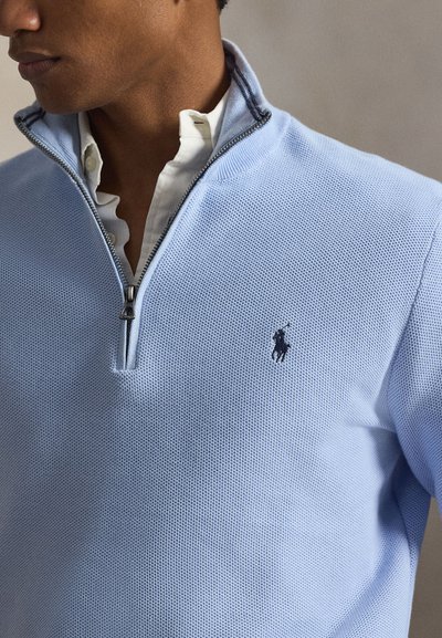 Mann trägt hellblauen, strukturierten Viertel-Zip-Pullover mit dunklem Logo, darüber ein weißes Hemd mit Knöpfen, schaut nach links.