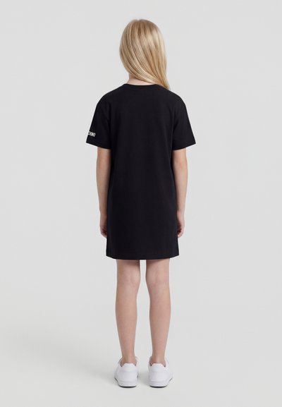 Otroku z dolgimi svetlimi lasmi, oblečenim v črno obleko s kratkimi rokavi in belimi supergami, stoji obrnjeno stran proti enobarvnemu ozadju.