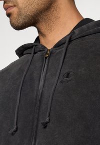 Κοντινό πλάνο ενός άνδρα που φοράει μαύρο φούτερ με φερμουάρ και κορδόνια, με διακριτικό λογότυπο Champion στο στήθος.