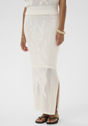 Jupe longue blanche en maille avec motif floral, fente latérale, portée avec des sandales noires et des bracelets beiges au poignet droit du mannequin.