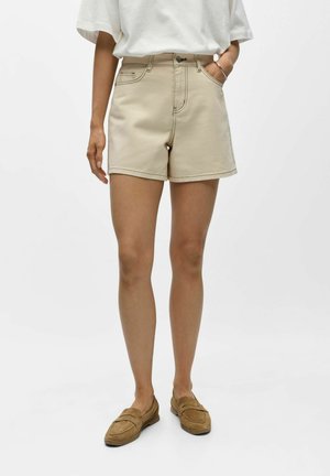 Kvinde iført beige shorts med høj talje, hvid løs t-shirt, brune ruskindsløbesko, med den ene hånd i lommen mod en ensfarvet baggrund.