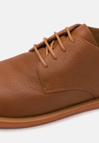 Scarpa in pelle marrone con superficie testurizzata, caratterizzata da dettagli in pizzo beige e una suola liscia color tan, progettata in uno stile a taglio basso.
