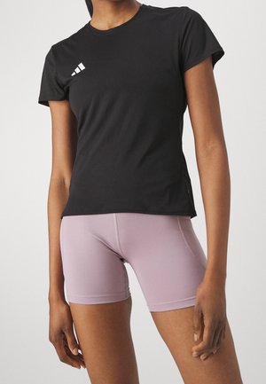 Femme portant un t-shirt de sport noir à manches courtes avec un logo et un short ajusté violet clair, debout sur fond blanc.