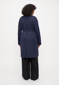 Marinblå trenchcoat med bälte i midjan, långa ärmar och veck bak. Tillverkad av slätt tyg med en medellång skärning.