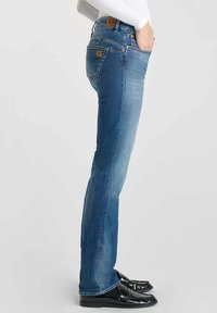 Jeans en denim bleu avec une coupe droite, texture délavée et patch en cuir marron à l'arrière. Associés à des chaussures noires.