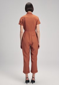 Mono de color óxido con cuello tipo camisa, mangas cortas, cintura ajustada y puños doblados. El material parece suave y ligeramente brillante.
