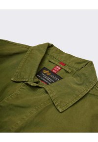 Camisa verde oliva con cuello, que presenta una etiqueta con la marca y la información de cuidado. Tela de algodón texturizado con costuras visibles.