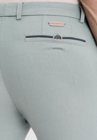 Hellgrüne Hose aus strukturiertem Stoff, schlanke Passform, Gesäßtasche mit Knopf und einem Markenlabel über der Tasche.